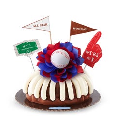A Chocolate Chocolate Chip bundt cake with frosting, decorated with a baseball on top of a purple and white bow in the center of the cake. The cake also contains a We're #1 foam finger, a flag that says All Star and a card that says M.V.P.